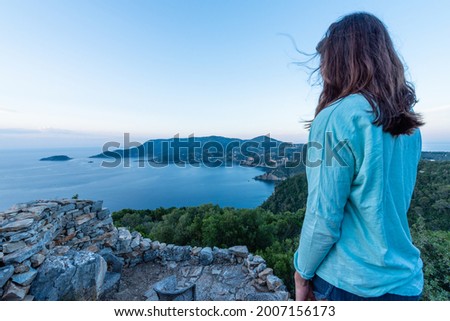 Woman on her back standing over the coastline landscape at dawn. She is wearing a light blue shirt. She is surrounded by a wild nature and clear sky
