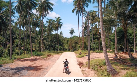 Woman On Motorbike Driving Through Palm Tree Lined Street On A Tropical Island.