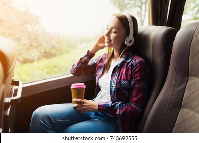 The woman on the passenger seat of the bus listens to music and drinks coffee. She looks out the window and smiles. Outside the window is a beautiful green landscape.