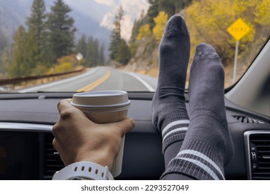 Woman on a passenger seat holding her feet and a cup of coffee on the dash, while travelling throught the fall mountains
