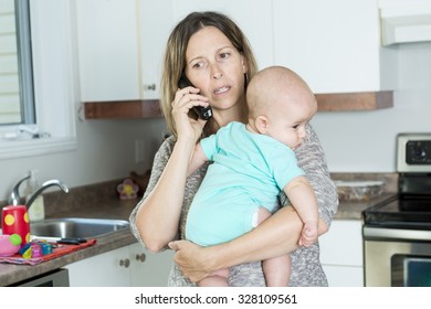 A Woman On The Phone While Holding Her Baby In Her Arms In The Kitchen