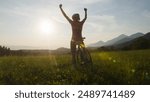 Woman on a road racing bike stopping at a green glade, celebrating with arms raised while looking at an amazing mountain panorama at sunset, handheld shot.