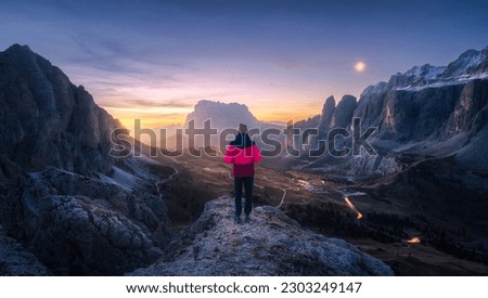 Woman on the rock and mountain peaks at night in autumn in Dolomites, Italy. Girl on the stone, high rocks, sky with moon, light trails on road at twilight in fall. Colorful landscape with cliffs
