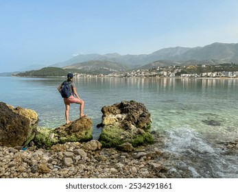 Woman on rock with panoramic view of coastline of tourist town Himare, Vlore, Albania. Sand beach Potami with scenic vistas of majestic Ceraunian mountain range. Summer vacation at Mediterranean sea