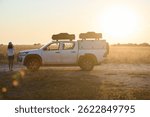 Woman on safari. Exploring African Savannah  stanfing next to an off-road 4x4 in Botswana’s wilderness. Ideal for: travel marketing, overlanding, nature exploration. Colours: gold, beige, white.