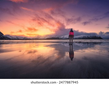 Woman on the sandy beach, snowy mountains and colorful sky at sunset in winter. Lofoten islands, Norway. Landscape with silhouette of a girl, sea, reflection in water, purple sky with pink clouds - Powered by Shutterstock