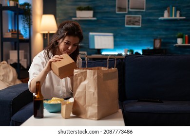 Woman On Sofa Unpacking Burger Box From Paper Takeaway Bag Sitting On Couch In Front Of Table With Beer Bottle, Fries And Bowl Of Chips. Person In Living Room Unpacking Fast-food Home Delivered.