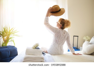 Woman On Vacation Sitting On A Bed In A Hotel Room With Hat In Hand In Front Of A Window.