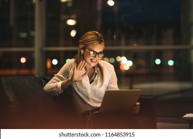 Woman on video call using a digital tablet sitting in office lobby. Smiling businesswoman late at night in office making gestures on a video call.