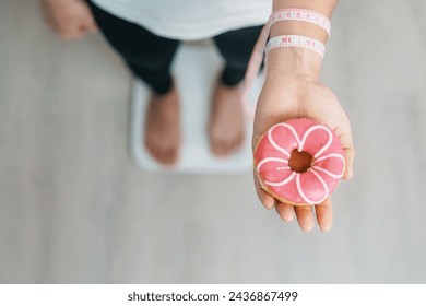 woman on weight scale and hand hold pink Donut, choose stop eating sweet is Unhealthy ealthy food. Dieting control, Weight loss, Obesity, eating lifestyle and nutrition concepts