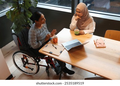 Woman on wheelchair and woman wearing hijab working at table in modern office - Powered by Shutterstock