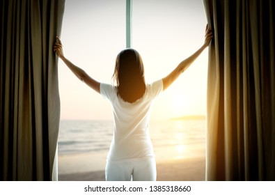 The woman is on the window in the bedroom. She opened the curtain at the window. In the morning and she looked at the view of the sea beach during the sunrise time