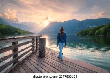 Woman on the wooden pier, mountains and Bohinj lake at sunset in spring. Girl, lake with reflection in water, green forest on mountains and colorful sky in summer. Triglav national park in Slovenia - Powered by Shutterstock