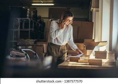 Woman online seller confirming orders from customer on the phone. E-commerce business owner looking at the papers and talking on phone in store warehouse. - Shutterstock ID 1387577570
