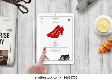 Woman online shopping red shoes with tablet. Device on table with newspaper, scissors, coffee, camera, croissant.