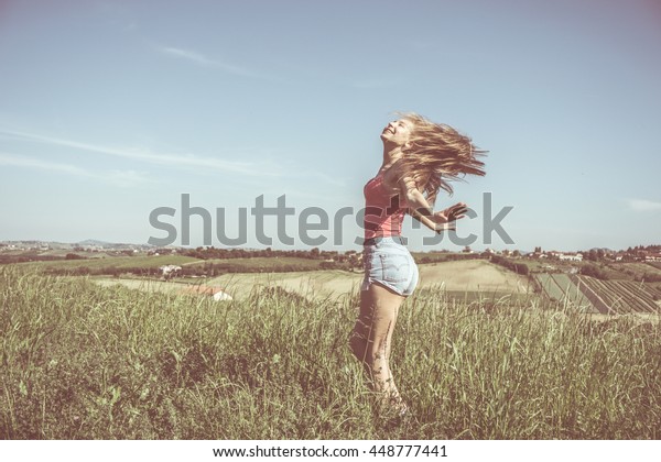 woman with open arms and long blond hair enjoying\
freedom in rural panoramic field of grass in summer - concept of\
wellness serenity breathing fresh air - vintage style faded colors\
and haze added