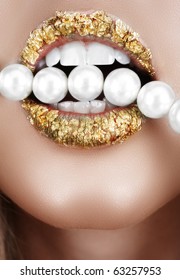 Woman open mouth with gold leaf make-up and teeth biting on faux pearls.