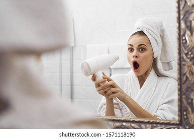 Woman With Open Mouth Holds A Hairdryer In Her Hand And Looks At Herself In The Mirror