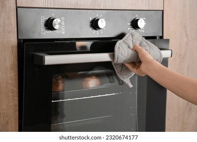 Woman opening electric oven with buns in kitchen, closeup