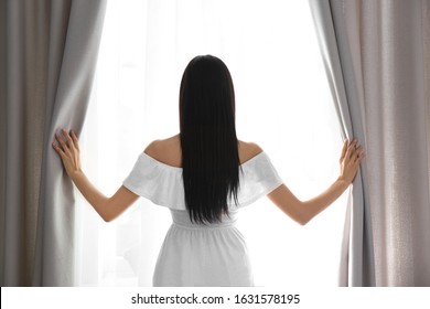 Woman Opening Window Curtains At Home In Morning, Back View