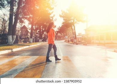 A Woman In An Orange Jacket, Walking Alone On A Pedestrian Crossing. In The Background, An Empty Road And Street. After The Rain. In Profile View. Light And Copy Space