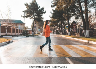 A Woman In An Orange Jacket, Walking Alone On A Pedestrian Crossing. In The Background, An Empty Road And Street. After The Rain. In Profile View. Light