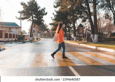 A Woman In An Orange Jacket, Walking Alone On A Pedestrian Crossing. In The Background, An Empty Road And Street. In Profile View. After The Rain