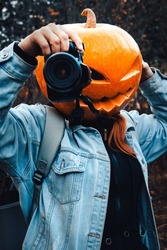 Woman With An Orange Pumpkin On His Head In A Jeans Jacket Stands On The Autumn Dry Grass. Scarecrow Takes Photos With A Camera. Concept Of Celebrating Halloween.