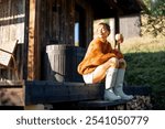 A woman in an orange sweater and boots sits on a wooden porch with a warm drink, enjoying the fresh morning air. Sunlight gently illuminates the cozy scene