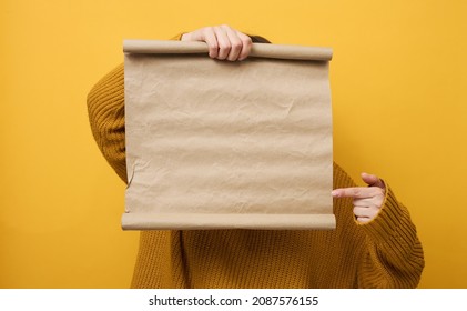 Woman In Orange Sweater Holds Blank Brown Brown Kraft Paper Roll. Place For An Inscription, Advertisement, Information