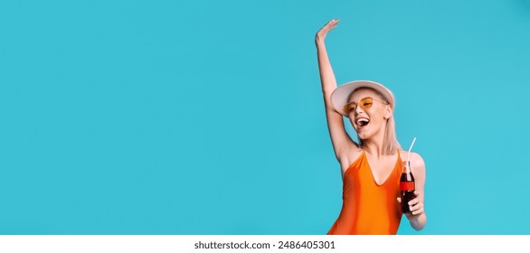 A woman in an orange swimsuit smiles and holds a soda while wearing a white sun hat and yellow sunglasses. She stands against a solid blue background with her arm raised, copy space - Powered by Shutterstock