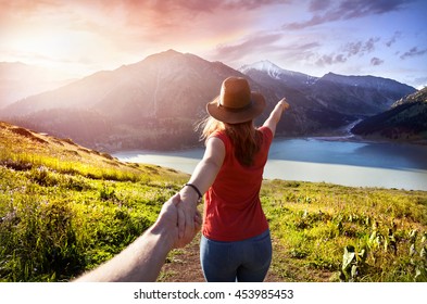 Woman in orange t-shirt and hat holding man by hand and going to the lake in the mountains at glowing sunrise 