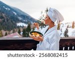 Woman ordered breakfast in hotel room eating food on balcony wearing robe enjoying winter mountains landscape. Vacation in Carpathians