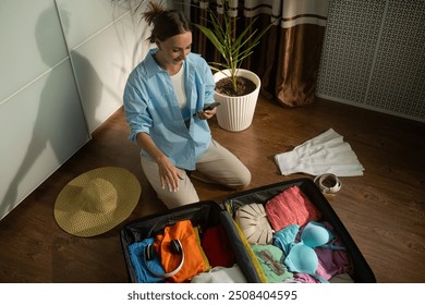 Woman organizes clothes and a sun hat while checking a packing list on her smartphone. Carefully placing items into a suitcase, she prepares for an upcoming vacation - Powered by Shutterstock