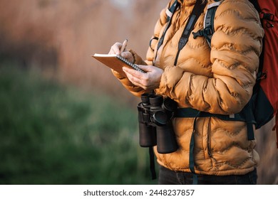 Woman ornithologist with binoculars writes details of bird behavior, migration and nesting sites in nature in a notebook. Bird watching outdoors
