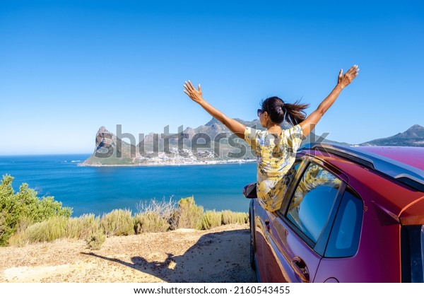 woman outside car window road trip with hands up, car at\
Chapmans Peak Drive in Cape Town South Africa looking out over the\
ocean. 