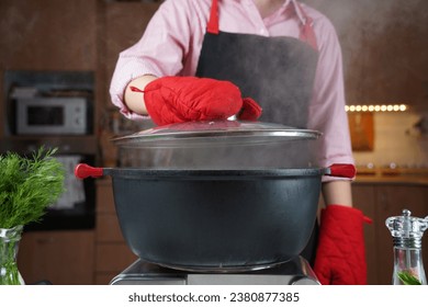 Woman  in oven heat gloves open lid of black pot with boiling water . Steam over pot