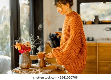 A woman in an oversized cozy orange sweater prepares pour-over coffee, with warm sunlight filling the rustic kitchen - Powered by Shutterstock