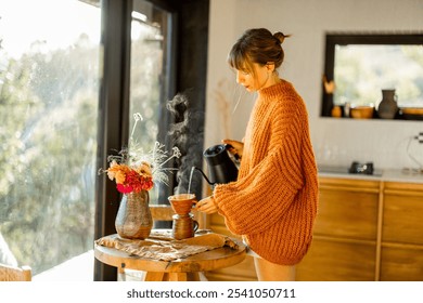 A woman in an oversized cozy orange sweater prepares pour-over coffee, with warm sunlight filling the rustic kitchen - Powered by Shutterstock