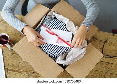 Woman Packaging A Christmas Gift For Posting Tying A Red Ribbon Around An Envelope On A Giftwrapped Box In A Cardboard Carton, High Angle View