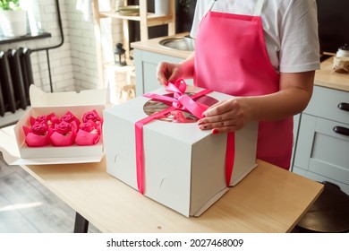 Woman Is Packing Big Birthday Cake Into A Box, Pink Cupcakes In The Package On The Box. Desserts Delivery Concept. 