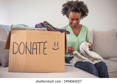 Woman Packing Clothes Into Donation Box In Living Room. Girl Puts In A Box With Donations Items. Volunteering. Woman Participating At Charity And Holding Donation Box