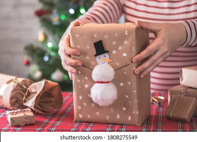 Woman Packing Handmade Christmas Gifts For Family Sitting At Table With Christmas Tree With Toys And A Glowing Garland With Bokeh. DIY. 