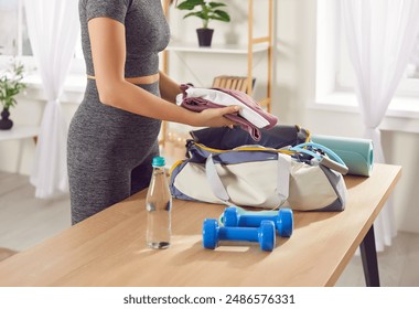 Woman is packing sports bag with essential items for fitness while going to sports training in gym. Woman puts sports uniform in bag, next to which lies pair of blue dumbbells and bottle of water. - Powered by Shutterstock