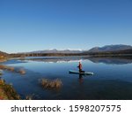 A woman paddle boarding on an Alaska lake in the fall. The lake is beginning to freeze close to the shore. The mirror smooth lake reflects the mountain range and the bight blue sky. 