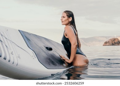 Woman Paddleboard Water Sports - Woman in a black swimsuit sits on a paddleboard in the water. The paddleboard is white and has a black pad on top. The woman is looking to the right, and the water is - Powered by Shutterstock