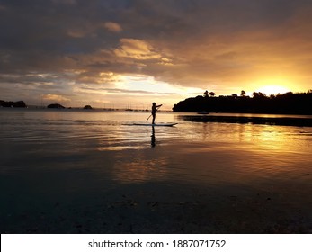 A woman is paddleboarding on a quiet beach at sunset, with orange colours and some boats on the back. This photo was taken in Shelly Beach, Waiheke Island, Auckland Region, New Zealand. January 2018 