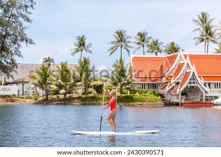 Woman paddleboarding on tranquil lake with tropical resort backdrop. Vacation and leisure.