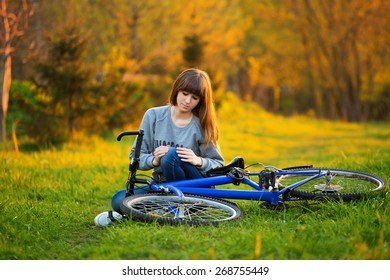 Woman With Pain In Knee Joints After Biking On Bicycle In Park At Sunset. Girl Sitting Down With Painful Face Expression. Knee Pain Bike Injury.