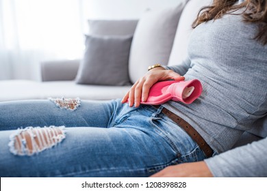 Woman in painful expression holding hot water bottle against belly suffering menstrual period pain, lying sad on home bed, having tummy cramp in female health concept